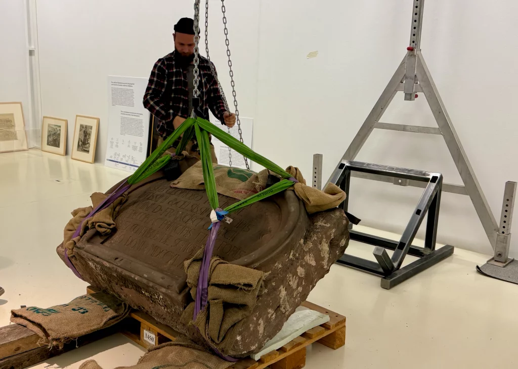 Eine restaurierte Steintafel, wird mithilfe eines Krans für die Ausstellung zurecht positioniert.
