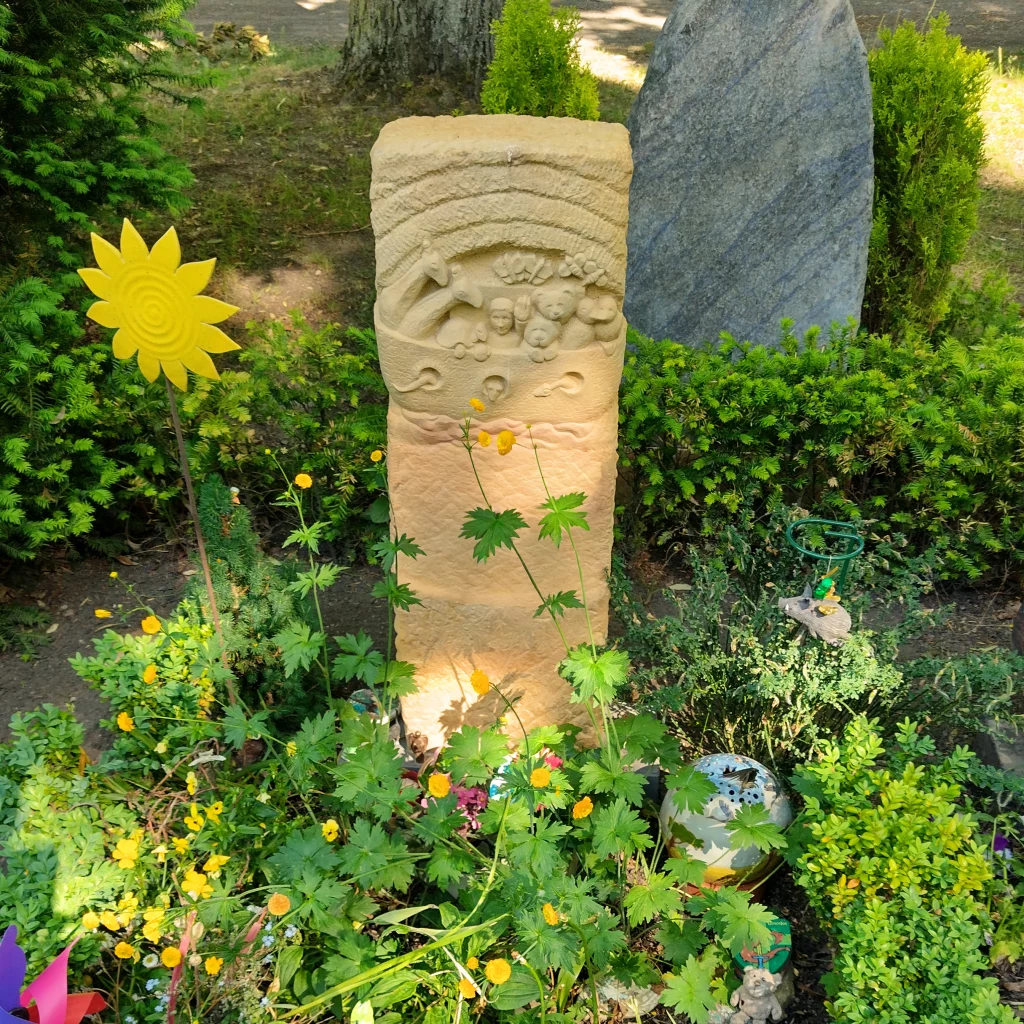 Ein Grabstein mit dem Motiv der Arche, welcher umringt ist von Blumen.